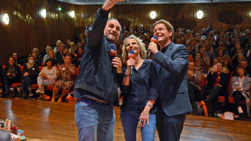Die drei Hosts Daniel Kaiser (von links), Katharina Mahrenholtz und Jan Ehlert blicken im Stehen glücklich auf ein Smartphone und machen ein "Selfie" von sich und dem Publikum beim Fanfest zur 100. Folge von eat.READ.sleep in Hannover