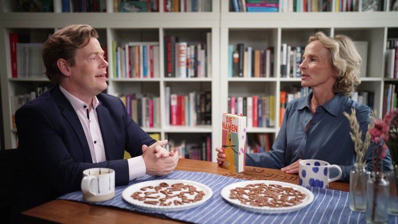 Jan und Katharina sitzen an einem Tisch, auf dem eine blau-weiß gestreife Decke liegt und zwei Teller mit Buchstabenkeksen stehen. Katharina hält das Buch "Die Namen" in der Hand.