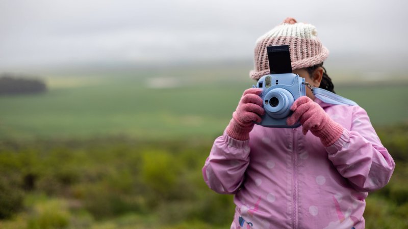 Ein Mädchen steht vor einer Landschaft und hält eine Polaroidkamera vors Gesicht