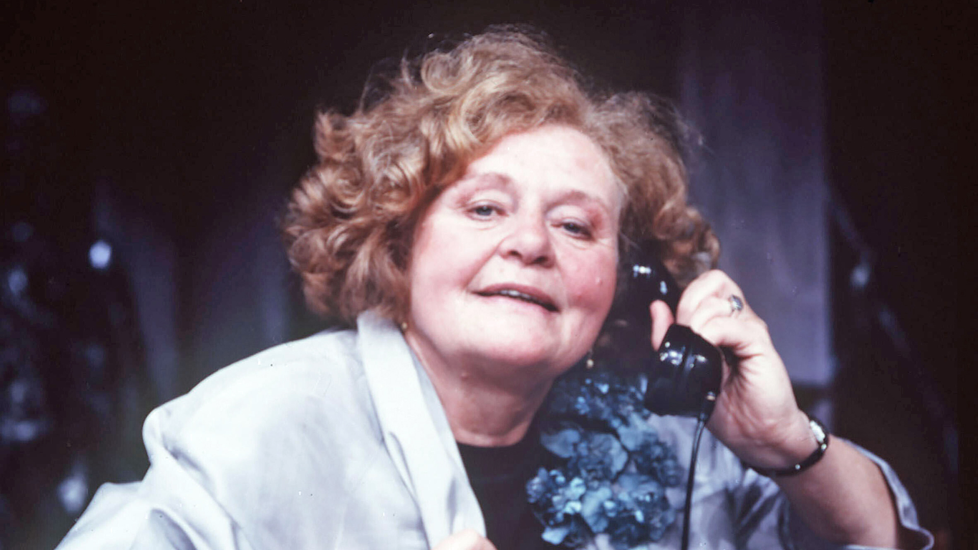 Ein Frau guckt in die Kamera und hat ein altes Telefon am Ohr.