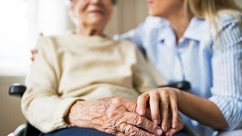 Eine Pflegerin legt den Arm um eine alte Frau im Rollstuhl und hält deren Hand.