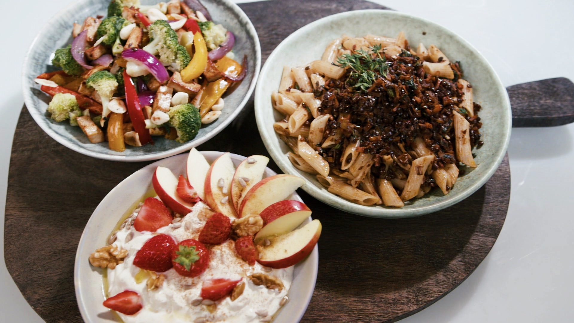 Auf einer Holzplatte stehen drei Schüsseln: Quarkcreme mit Früchten, Brokkoli-Tofu-Wok und Penne mit Balsamico-Linsen-Bolognese.