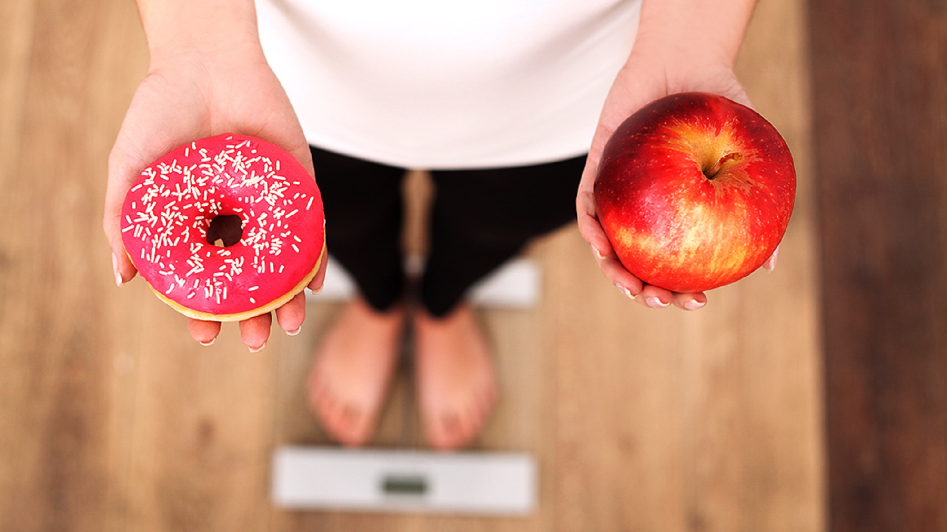Eine Frau steht auf einer Waage und hält einen Donut und einen Apfel in den Händen.