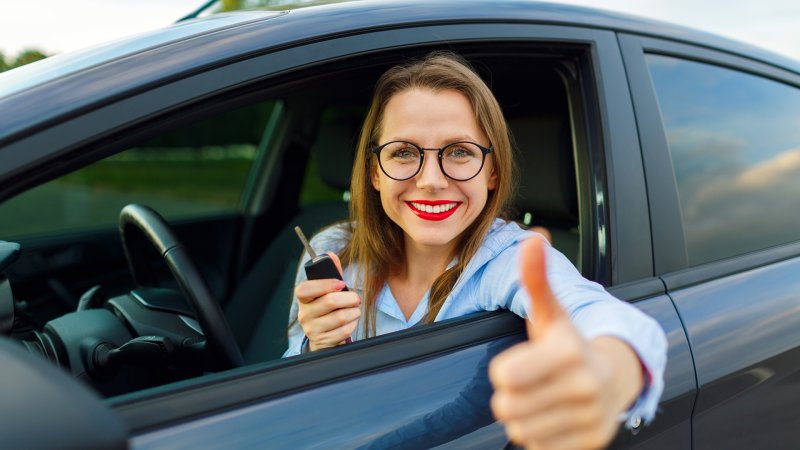 Eine junge Frau sitzt im Auto und hält einen Autoschlüssel in der Hand, sie streckt ihren Daumen hoch.