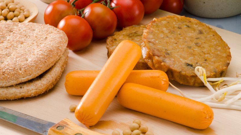 Fleischlose Würstchen und Bratlinge liegen auf einem Brett neben Tomaten und Brot.