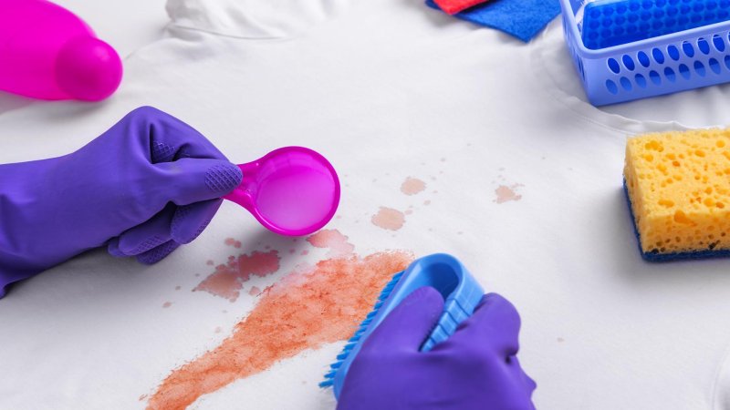 Hände mit Gummihandschuhen, Bürste und Dosierlöffel über einem weißen T-Shirt mit Fleck
