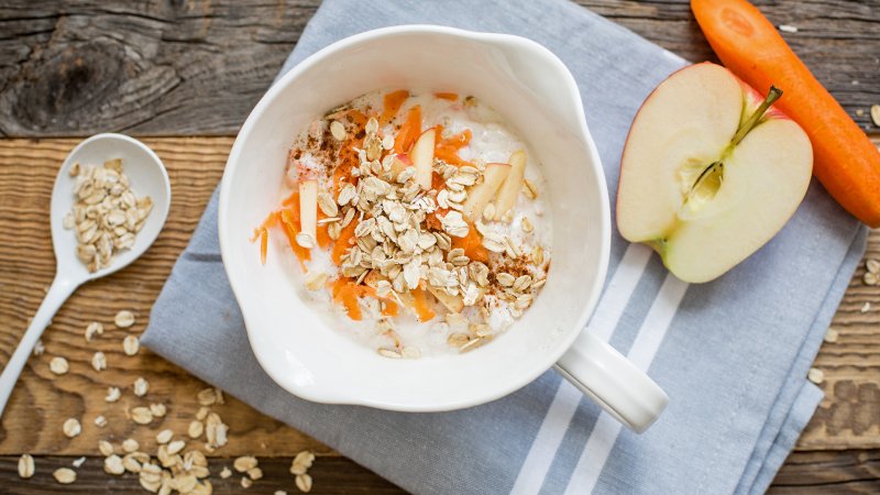 Auf einem Holztisch steht ein Schälchen Hafer-Frühstück, daneben ein halber Apfel sowie eine Möhre.