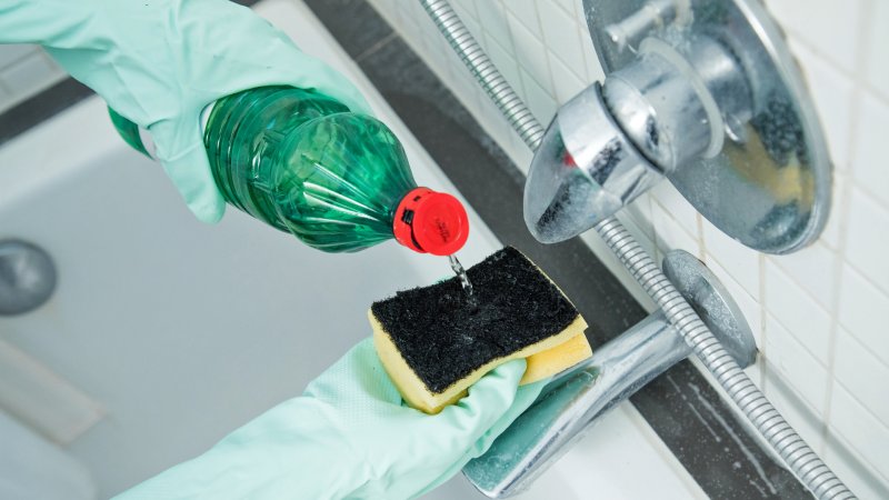 Hände mit Gummihandschuhen mit Schwamm und Essigflasche vor verkalkter Badewannenarmatur
